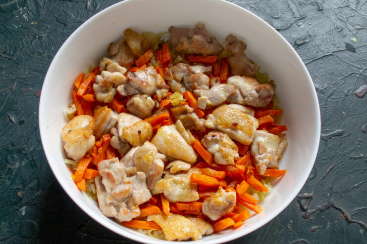 Adding the layer of browned chicken over the vegetables for the plov. Golden-brown chicken pieces being layered on top of sautéed onions and carrots in a baking dish.