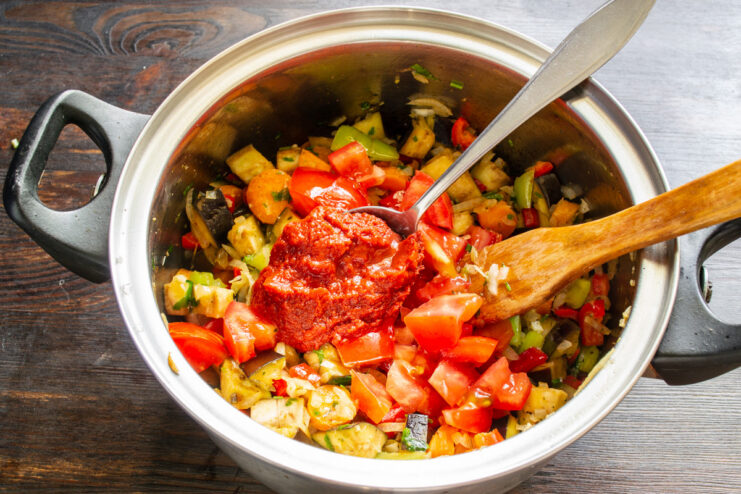Adding tomato paste to deepen the flavor and color of the ratatouille sauce. A spoonful of rich tomato paste being added to the vegetable stew in a pot.