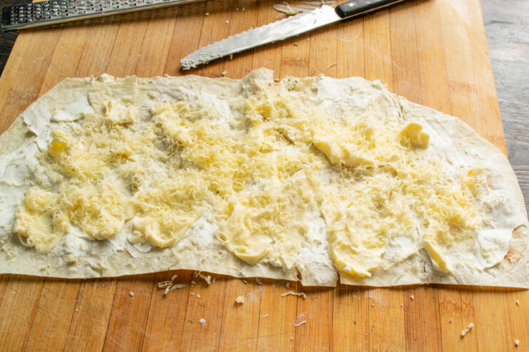 Adding the final layer of grated hard cheese Sprinkling freshly grated hard cheese over the layers of processed and cream cheese on lavash.