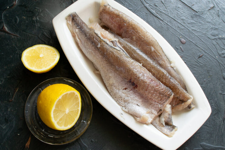 Raw hake fillets in a glass dish being drizzled with fresh lemon juice.