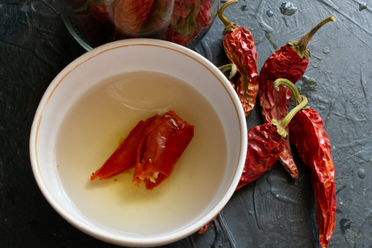 A hand holding a fresh red chili pepper next to a bowl of dried chili peppers.