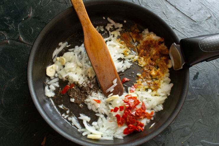 Spices, including paprika and chopped chili, being added to a pan with sautéing onions and garlic.