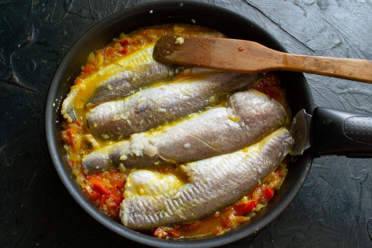 Marinated hake fillets being carefully placed into a pan of simmering red vegetable sauce.