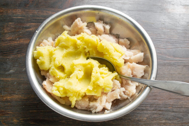 Combining fish mince and cooled mashed potatoes to make the patty mixture. A bowl with minced fish and a portion of mashed potatoes being added to it.