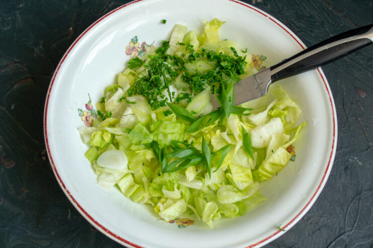 Adding fresh herbs to the salad for extra flavor and aroma. Chopped parsley and green onions being added to a bowl of shredded iceberg lettuce.