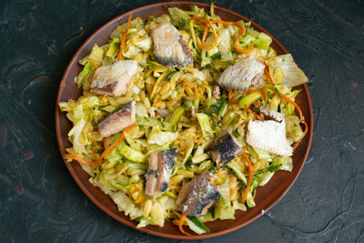 Adding the star ingredient: tender canned saury. Pieces of canned saury fish being placed on top of the pasta and vegetable salad.