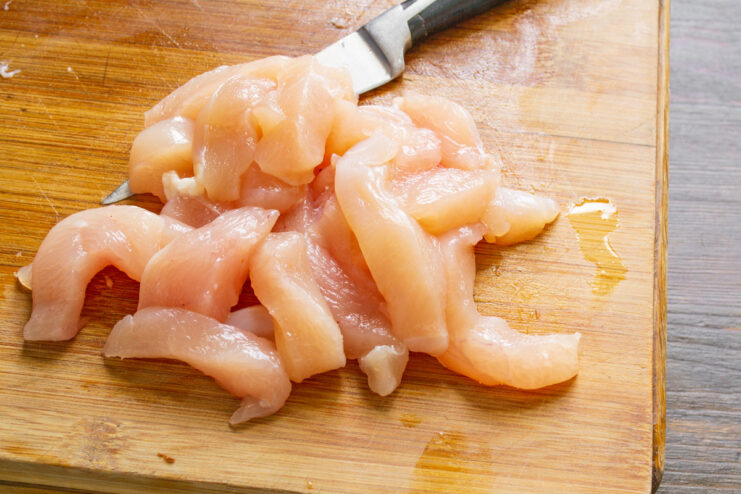 Slicing chicken breast against the grain ensures tender, juicy pieces for the salad. Raw skinless chicken breast being cut into even strips on a cutting board.