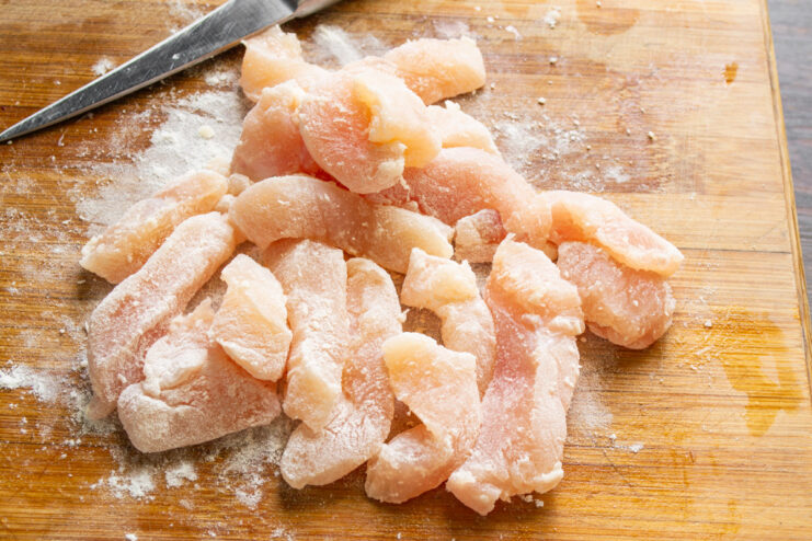 Coating the chicken in potato starch creates a light, crispy crust when fried. Sliced raw chicken in a bowl being tossed with white potato starch.
