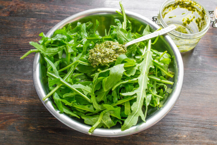 Adding pesto sauce directly to the arugula infuses every leaf with flavor. A bowl of fresh arugula with a dollop of green pesto sauce on top.