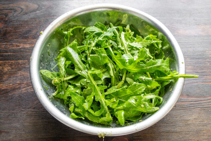 Tossing the arugula ensures it's evenly coated with the delicious pesto dressing. Arugula leaves being tossed with pesto sauce in a large mixing bowl.