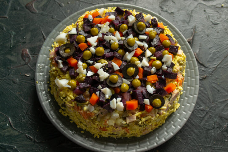 Final touches: arranging the colorful vegetable garnish on top of the salad. Adding the final garnish of chopped purple carrot, olives, and peas to the Stolichny Salad.