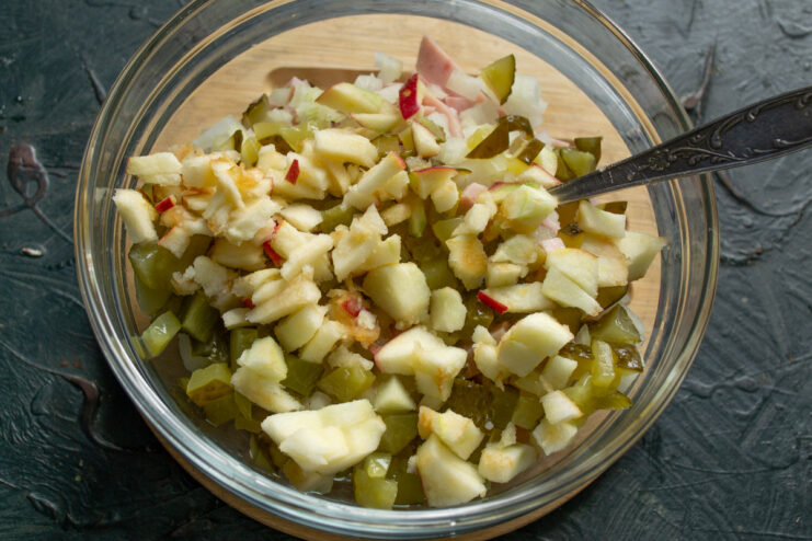 Chopping dill pickles and a tart-sweet apple to add crunch and flavor. Finely chopped dill pickles and a tart-sweet apple on a cutting board.