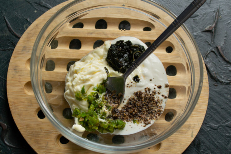 Preparing the creamy and spicy dressing for the Stolichny Salad. A bowl with mayonnaise, smetana, and green adjika being mixed for the salad dressing.