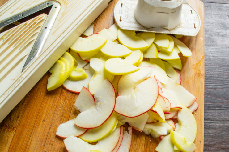 A hand using a mandoline slicer to cut a peeled apple into paper-thin slices for a pie.