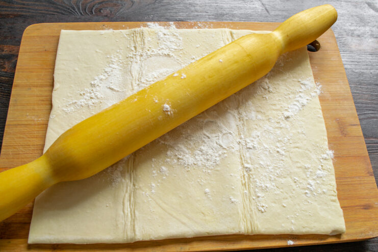 Rolling out a sheet of puff pastry with a wooden rolling pin on a floured board.