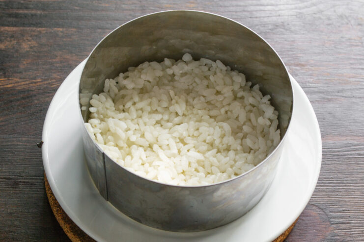 Forming the Rice Layer in a Cooking Ring Pressing cooked rice into a metal cooking ring on a plate to form the first layer of the salad.