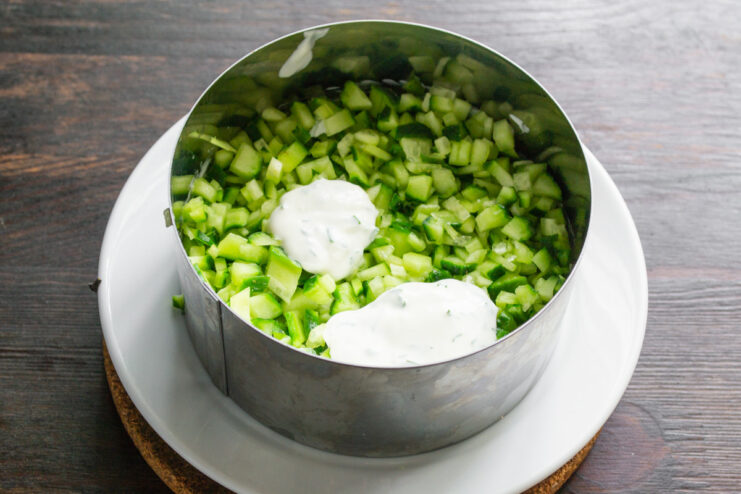 Adding the Creamy Dressing to the Cucumber Layer The cucumber layer in the cooking ring being covered with a creamy white dressing.