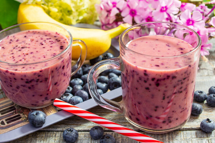 A glass of purple banana and blueberry smoothie, garnished with fresh blueberries and a straw.