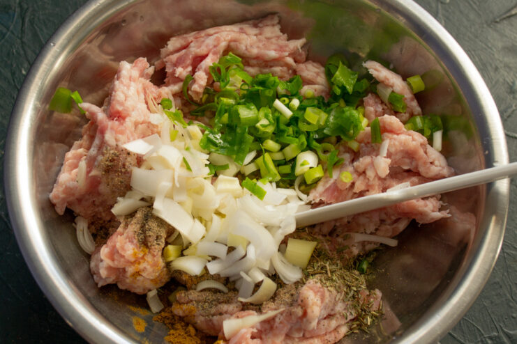 Adding finely chopped leeks and green onions to the meat mixture. Finely chopped leeks and green onions being added to a bowl of ground meat.