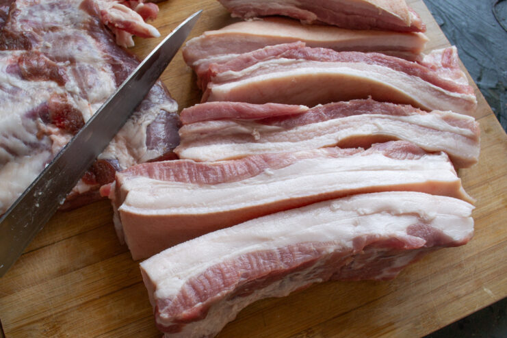 Step 1: Slicing the Pork Belly for Curing Raw pork belly being cut into long, thick strips on a wooden cutting board.