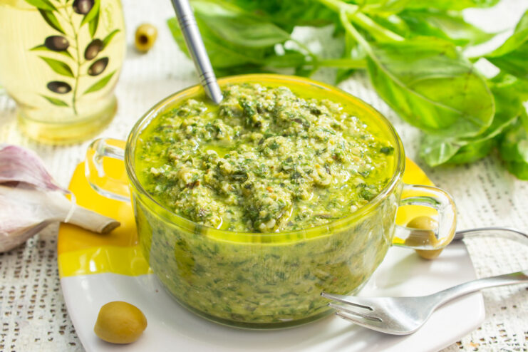 A glass jar of green pesto sauce with olives, served with bread and fresh basil.