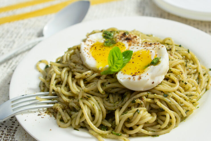 A white plate with a serving of spaghetti with green pesto sauce, topped with a halved soft-boiled egg and a basil leaf.