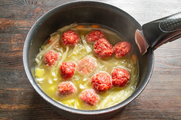 Adding the raw meatballs to the simmering vegetable soup Carefully placing raw meatballs into the pot of simmering vegetable soup.