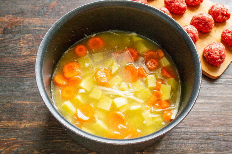 Adding broth to the vegetables to create the soup base Pouring broth into a pot with chopped vegetables like potatoes, carrots, and cabbage.