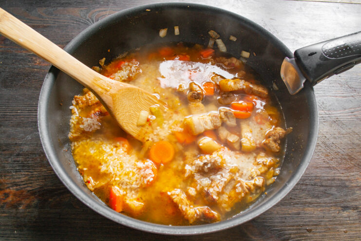 Adding Liquid to Cook the Rice Perfectly Hot water or broth being poured into the skillet over the rice, pork, and vegetables.