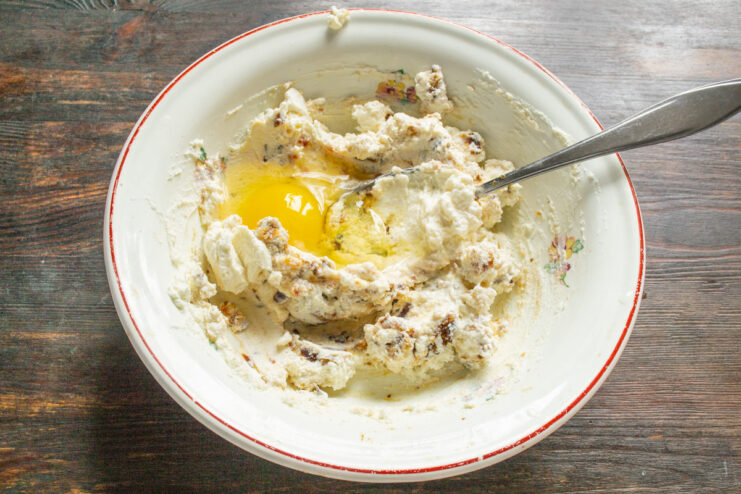 Adding an Egg to the Syrniki Mixture A large chicken egg being cracked into a bowl with ricotta and dates.