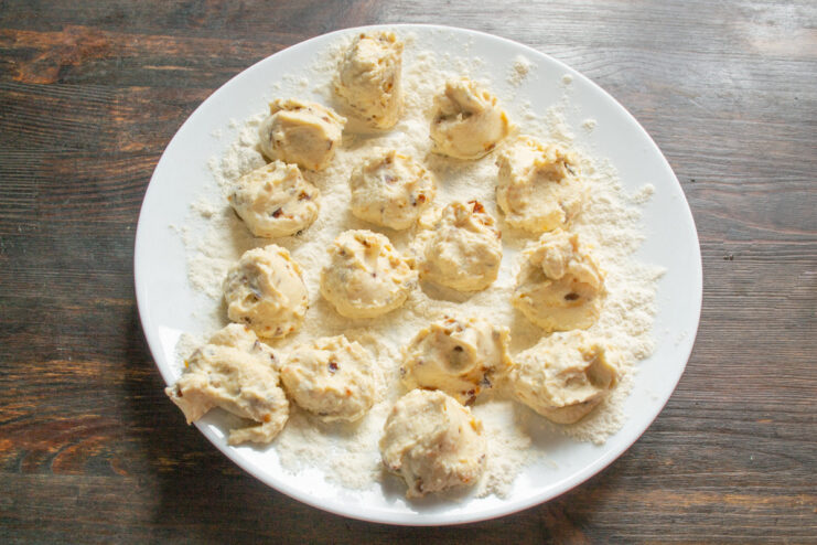Portioning the Syrniki Dough with a Teaspoon Scooping portions of syrniki dough with a teaspoon onto a plate of flour.