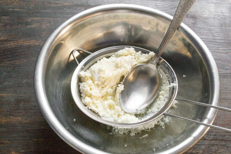 Sifting cottage cheese is the key to a smooth syrniki texture. White cottage cheese being pressed through a fine-mesh sieve into a glass bowl.