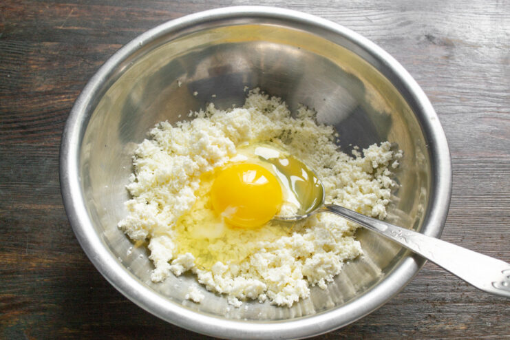 Adding an egg to bind the syrniki dough. An egg being cracked into a bowl of sifted cottage cheese.