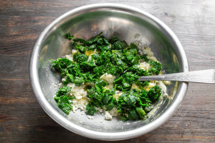 Incorporating finely chopped spinach into the syrniki mixture. Chopped green spinach being added to the cottage cheese and egg mixture in a bowl.