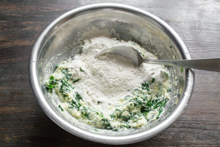 Adding flour to bind the syrniki dough. A spoonful of white flour being added to the cottage cheese and spinach mixture.