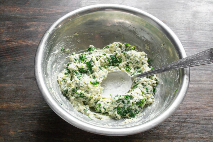 Mixing the syrniki dough until all ingredients are well combined. A spoon mixing the cottage cheese, spinach, and flour dough in a glass bowl.