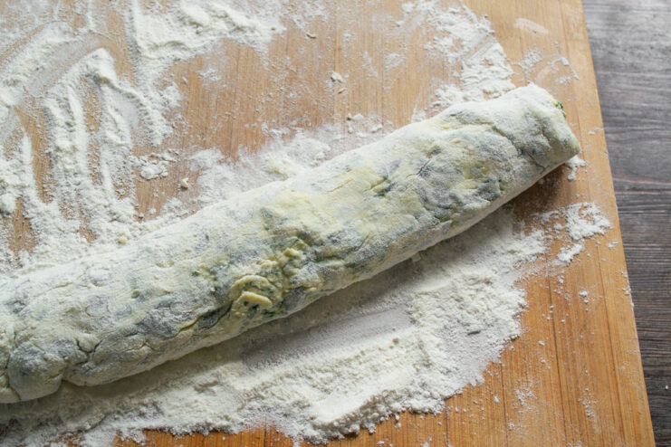 Shaping the dough into a log for easy portioning. Syrniki dough being rolled into a log on a floured wooden board.