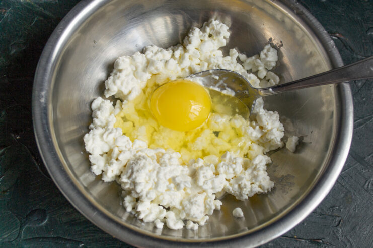 Adding an egg to the tvorog mixture A small chicken egg being cracked into the bowl with tvorog for the syrniki batter.