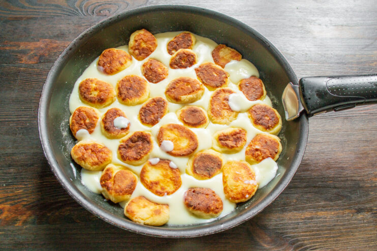 Finishing the dish: Pouring custard over the fried syrniki Pouring creamy yellow custard over freshly fried syrniki in a black skillet.