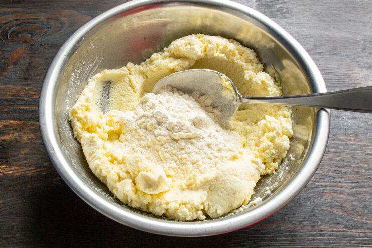 Step 2: Adding flour to the syrniki mixture Adding a spoonful of all-purpose flour to the cheese and egg mixture in a white bowl.