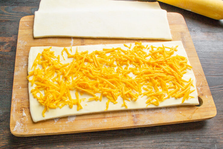 Adding a Generous Layer of Cheddar Cheese Grated cheddar cheese being spread over a sheet of egg-washed puff pastry.