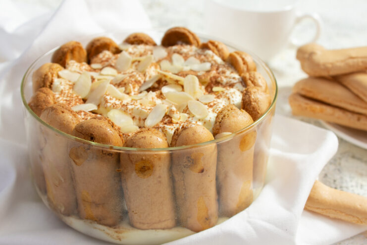 A whole eggless Tiramisu cake in a glass dish, dusted with cocoa and decorated with almond flakes.