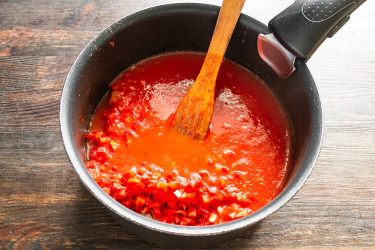 Combining the Tomato Purée with Sautéed Vegetables Smooth red tomato purée being poured into a saucepan containing diced onions and peppers.