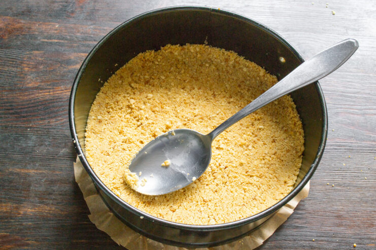 Step 4: Forming the No-Bake Cake Crust A spoon pressing down a cookie crumb mixture in a springform pan to create an even crust.