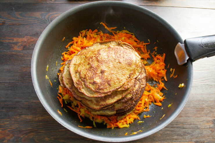 Layering cooked liver pancakes with sautéed vegetables to keep them moist A stack of cooked liver pancakes placed in a skillet on top of sautéed carrots and onions.