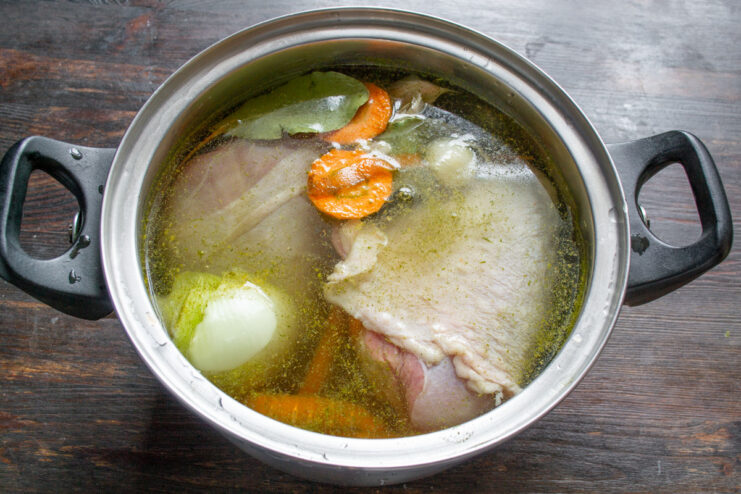 Starting the broth for the aspic with turkey drumsticks. Raw turkey drumsticks, an onion, and carrots in a large pot being covered with water to make broth.