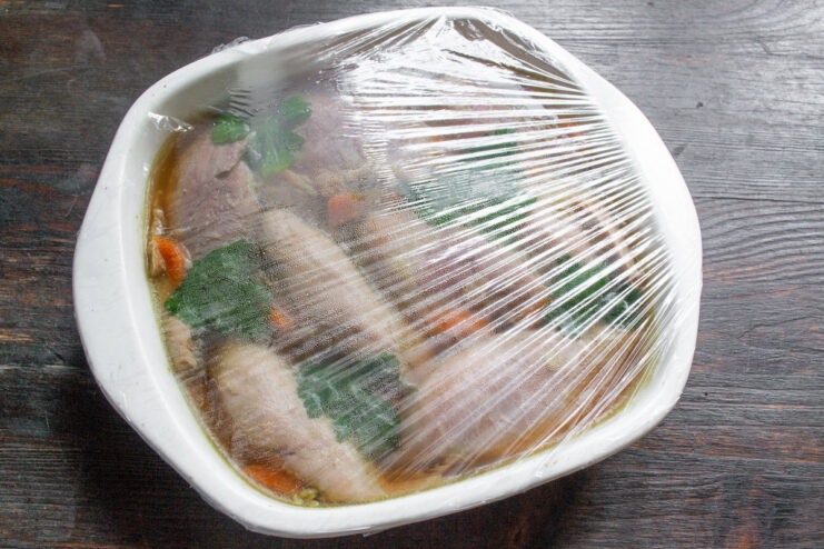 Preparing the aspic for chilling to let it set perfectly. A glass dish filled with turkey aspic mixture being covered tightly with plastic wrap.