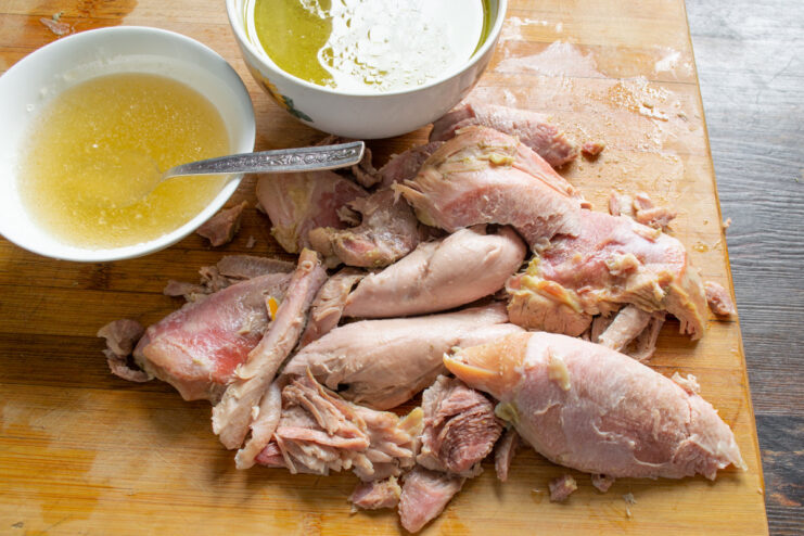 Preparing the tender turkey meat for the aspic. Cooked turkey meat being shredded into large pieces on a cutting board.