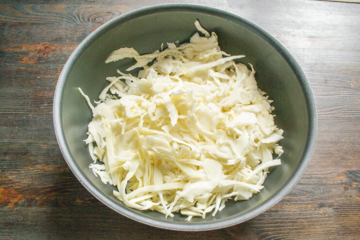 Shredding cabbage into thin strips for the pie filling. This ensures it cooks evenly and becomes tender. Hands shredding white cabbage into thin strips on a cutting board.