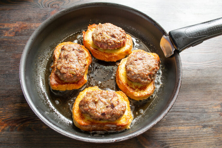 Assembling the Sandwiches in the Pan Placing a cooked beef patty on top of a slice of fried bread in the skillet.
